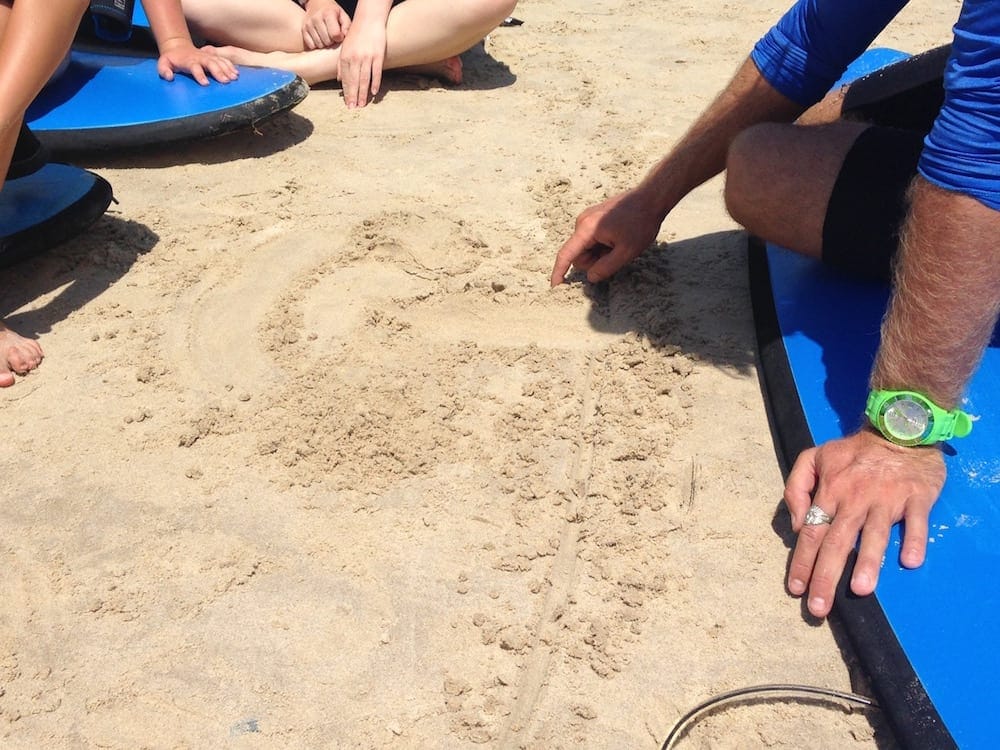Family Surfing Lessons Instructor draws instructions in sand on beach on a sunny day