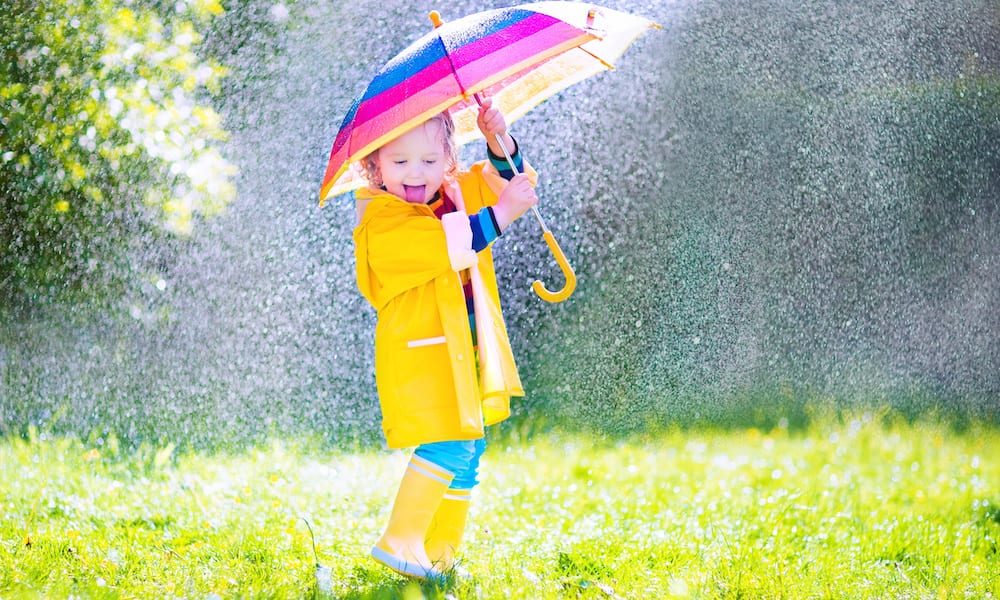 rainy day activities Funny cute curly toddler girl wearing yellow waterproof coat and boots holding colorful umbrella playing in the garden by rain and sun weather on a warm autumn or sumemr day