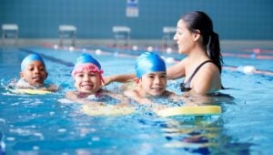 Female Coach In Water Giving Group Of Children Swimming Lesson In Indoor Pool