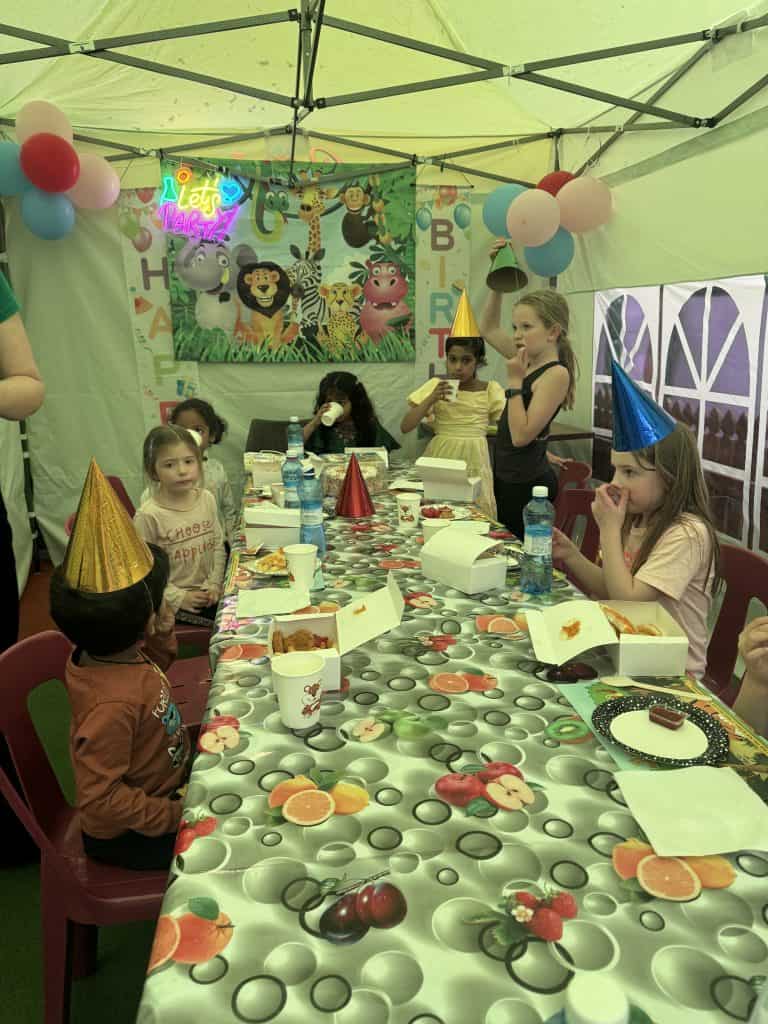 Children enjoying a colourful birthday party in Brisbane with decorations, party hats, and food under a marquee.