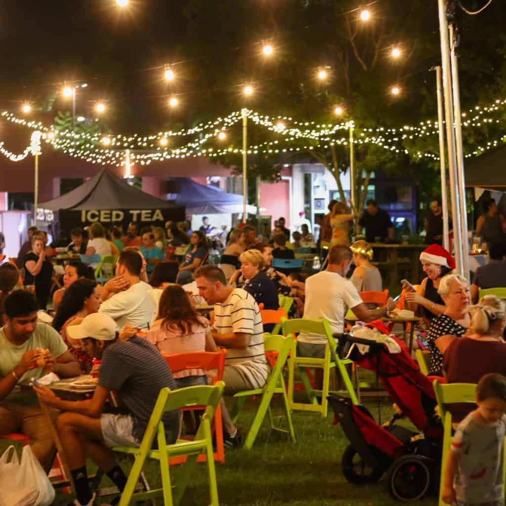 Families and friends enjoying the festive atmosphere at the Beenleigh Christmas Twilight Markets, seated under twinkling string lights with food stalls in the background. People chat, eat, and relax on colourful chairs while children play nearby, capturing the cheerful community vibe of the evening.