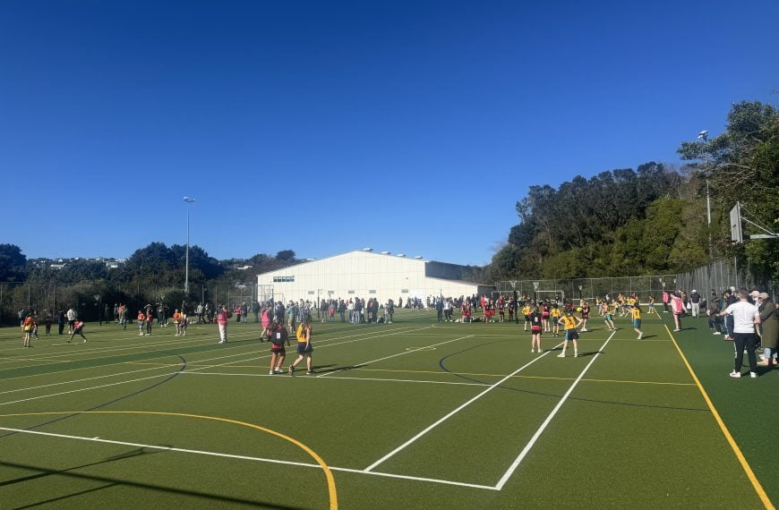 Junior netball players competing on outdoor courts with coaches, referees, and parents watching under a clear blue sky