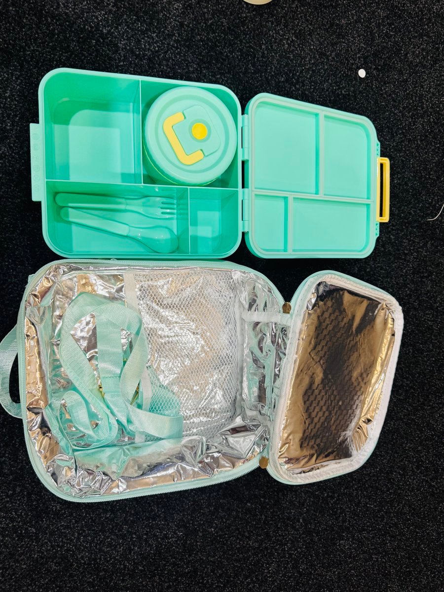 Open green bunny-print kids’ bento lunch box showing four compartments, stainless-steel soup cup, and matching fork and spoon beside its insulated lunch bag.