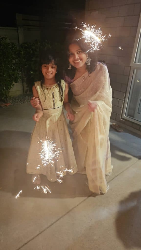 Mother and daughter celebrating Deepavali outdoors at night, smiling while holding sparklers and enjoying fireworks during the Festival of Lights.