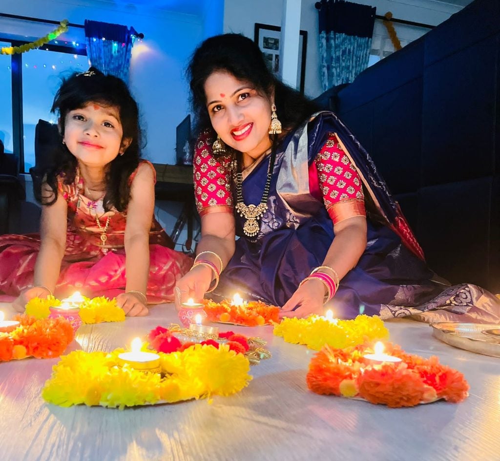 Mother and daughter celebrating Deepavali at home, lighting diyas surrounded by bright yellow and orange flower garlands during the Festival of Lights.