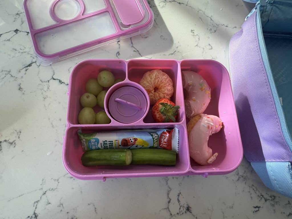 Our kidsโ lunch box filled with green grapes, cucumber sticks, a peeled mandarin, a strawberry, mini pink donuts, and a fruit strap