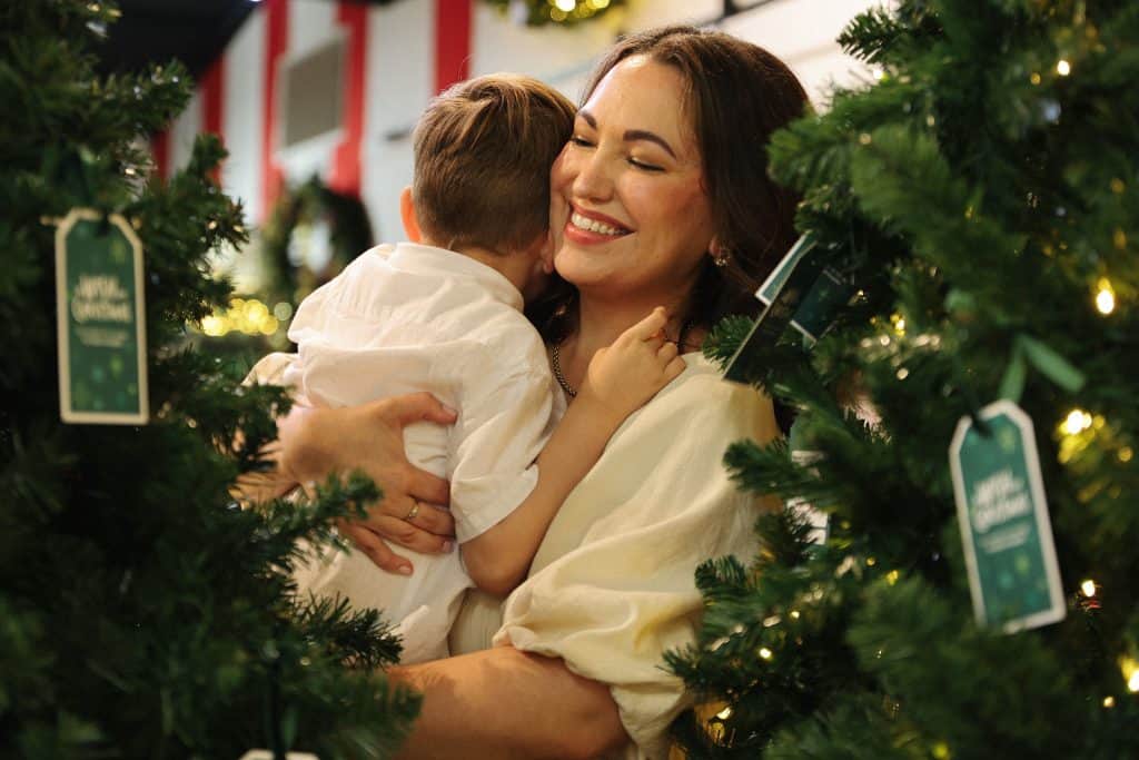 Mother hugging her young child among decorated Christmas trees, smiling warmly during a festive Childrenโs Hospital Foundation campaign.