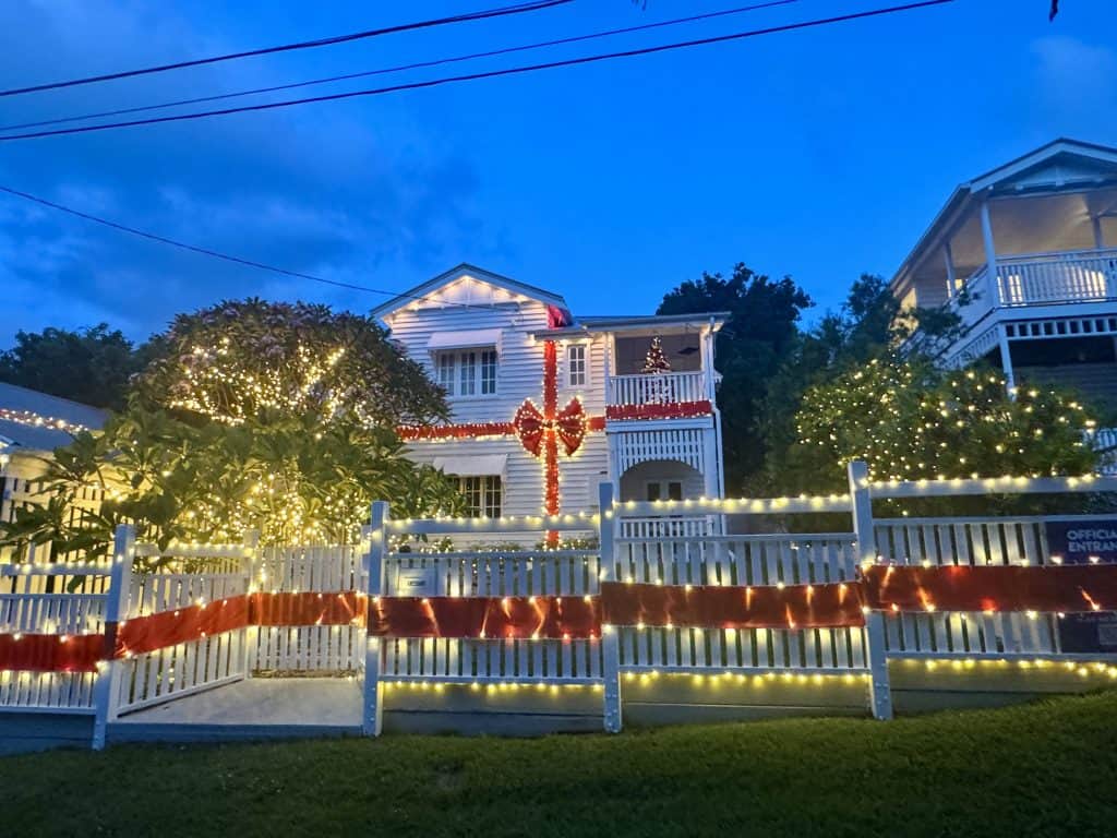 Christmas lights at 29 Billington Street in Brisbane featuring a white Queenslander home decorated with warm fairy lights, a large red bow, and festive tree on the balcony.